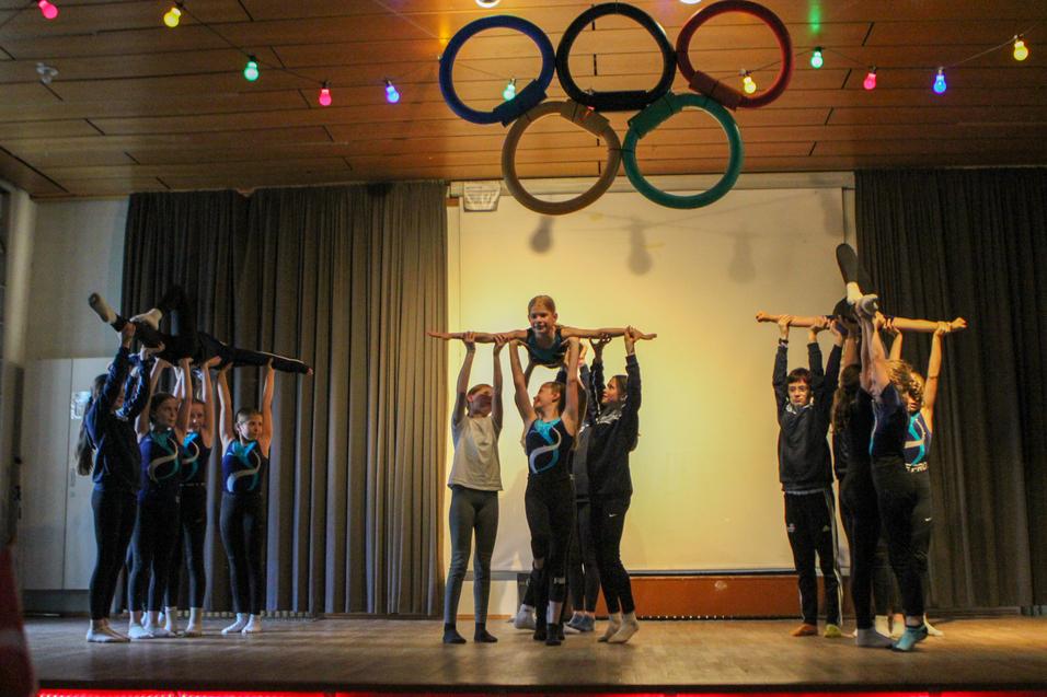 Gruppe von Turnerinnen führt eine Hebetechnik auf einer Bühne mit olympischen Ringen im Hintergrund aus.