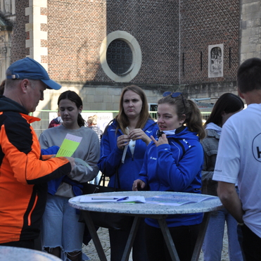 Eine Gruppe junger Menschen diskutiert mit einem Mann in orangefarbener Jacke vor einer historischen Mauer.