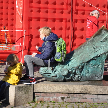 Junge mit Rucksack sitzt auf einem Brunnen mit Fischskulptur, während ein Kind in Gelb Eis isst. Im Hintergrund eine rote Wand.