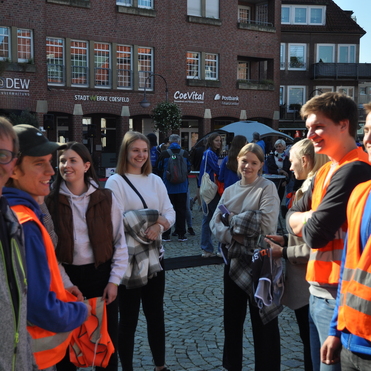 Gruppe junger Menschen, einige in orangefarbenen Westen, unterhalten sich auf einem Platz mit Häusern im Hintergrund.