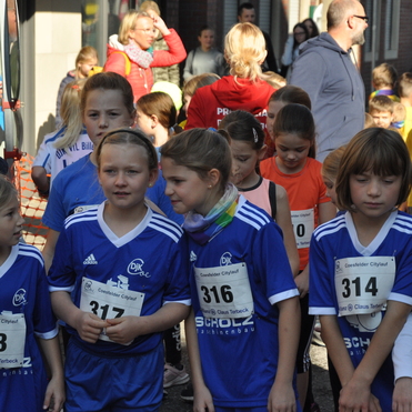 Gruppe junger Läufer in blauen Trikots mit Startnummern, bereit für einen Laufwettbewerb auf einer belebten Straße.