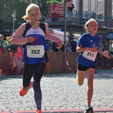 Zwei Mädchen in Sportkleidung laufen auf einer Stadtlaufstrecke über eine Zeitmessmatte. Zuschauer sind im Hintergrund sichtbar.