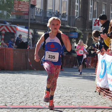 Mädchen in Sportkleidung läuft auf einem markierten Zielbereich eines Laufwettbewerbs, umgeben von Zuschauern.