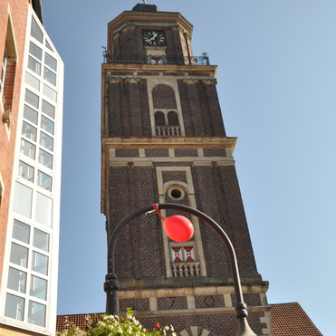 Stadtansicht mit einem hohen Backsteinturm, rotem Ball &uuml;ber einer Laterne und bl&uuml;henden Pflanzen im Vordergrund.