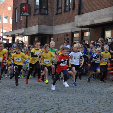 Kinder in Sportbekleidung starten gemeinsam einen Wettlauf auf einer Stra&szlig;e, Zuschauer applaudieren im Hintergrund.