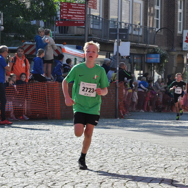 Ein Junge mit blondem Haar l&auml;uft in einem Wettlauf, umgeben von Zuschauern und einer roten Absperrung.