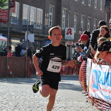 Ein junger L&auml;ufer in schwarzer Sportkleidung &uuml;berquert die Ziellinie eines Rennens vor jubelnden Zuschauern.
