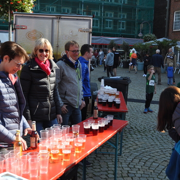 Personen stehen an einem Tisch mit Getr&auml;nkebecher in einer belebten Platzszene. Im Hintergrund sind Menschen unterwegs.