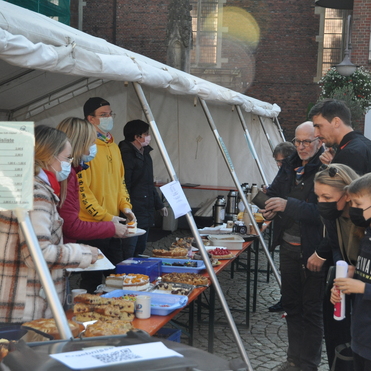 Verkaufstisch unter einem Zelt mit Menschen, die Kuchen und Getr&auml;nke an einem Marktstand ausw&auml;hlen.