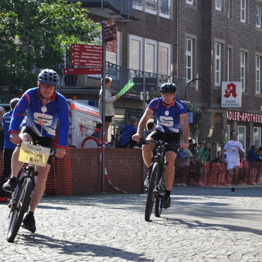 Zwei Radfahrer in blauer Sportkleidung fahren an einer Zuschauergruppe und einem roten Absperrgitter vorbei.