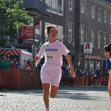 Ein junger L&auml;ufer in wei&szlig;em Trikot sprintet auf einer Stra&szlig;enszene zu einer jubelnden Menge.
