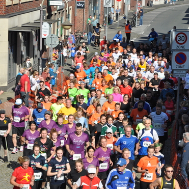 Teilnehmer eines Stra&szlig;enlaufs in bunten T-Shirts laufen an Zuschauern entlang auf einer st&auml;dtischen Stra&szlig;e.
