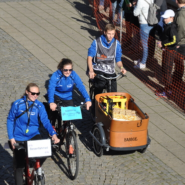 Drei Radfahrer in blauen Outfits fahren auf einer Pflasterstra&szlig;e, einer zieht einen Lastenanh&auml;nger mit Kisten.