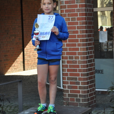 M&auml;dchen mit Pokal und Urkunde auf dem Siegertreppchen beim 27. Costfelder City-Lauf, umgeben von Ziegelwand.