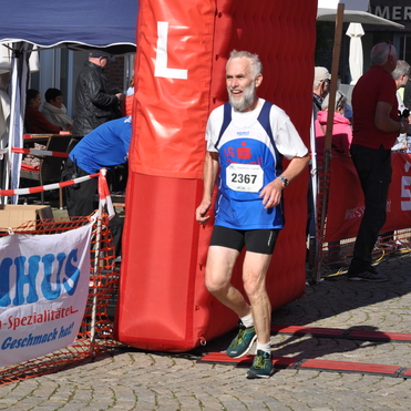&Auml;lterer m&auml;nnlicher L&auml;ufer mit Bart, der l&auml;chelnd das Zielband bei einem Wettlauf passiert. Im Hintergrund Menschen und Banner.