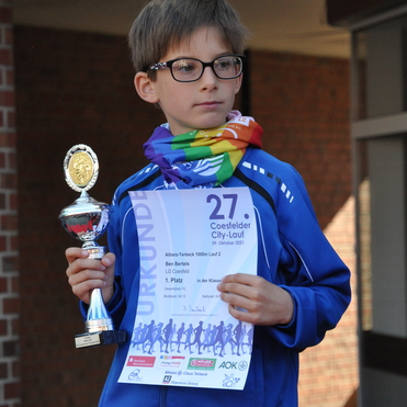 Junge mit Brille, Regenbogen-Schal, h&auml;lt Pokal und Urkunde f&uuml;r den 1. Platz beim Coesfelder City-Lauf 2021.