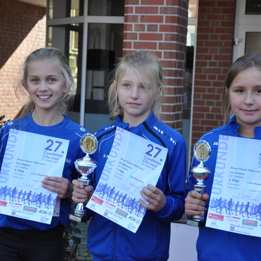 Drei M&auml;dchen in blauen Sportjacken halten Pokale und Urkunden eines Laufwettbewerbs, l&auml;chelnd vor einer Backsteinmauer.