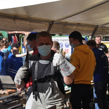 Grillender Mann in Arbeitskleidung mit Handschuhen und Grillzange, umgeben von Besuchern in einer Zeltatmosph&auml;re.