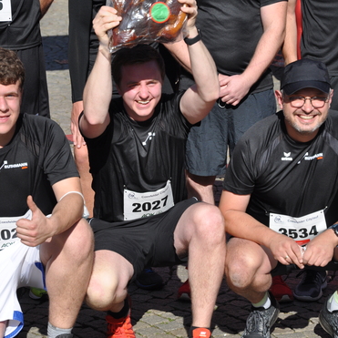 L&auml;ufer in schwarzen T-Shirts posieren nach einem Rennen, einer h&auml;lt eine gro&szlig;e Brezel in einer Plastikt&uuml;te &uuml;ber dem Kopf.