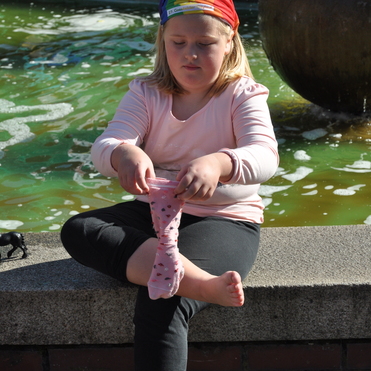 M&auml;dchen mit Regenbogenband sitzt am Brunnen und zieht einen rosa Socken mit Punkten an.