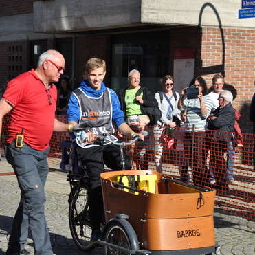 Ein junger Fahrer auf einem Lastenrad l&auml;chelt, w&auml;hrend ein Mann in rot ihm zur Seite steht; Zuschauer beobachten die Szene.
