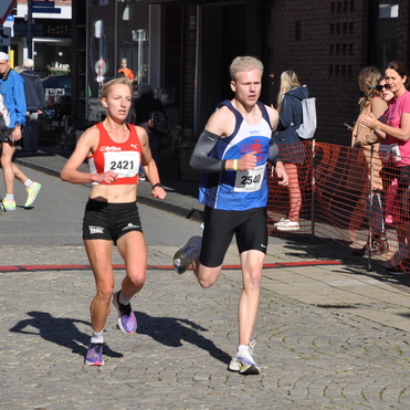 Zwei L&auml;ufer in Sportbekleidung &uuml;berqueren einen gepflasterten Weg, w&auml;hrend Zuschauer am Rand zuschauen und jubeln.