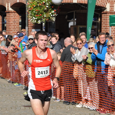 L&auml;ufer in roter Sportbekleidung am Ziel, umgeben von jubelndem Publikum und einer roten Absperrung.