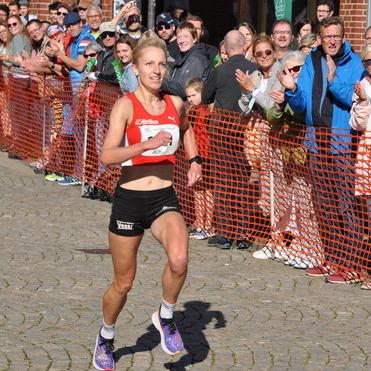 L&auml;uferin in rotem Shirt sprintet auf einer Veranstaltung, umgeben von jubelnden Zuschauern hinter einem orangefarbenen Absperrband.