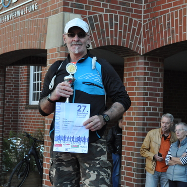 Mann in Sportkleidung h&auml;lt Pokal und Urkunde des 27. Coesfelder City-Laufs vor einem Backsteingeb&auml;ude.