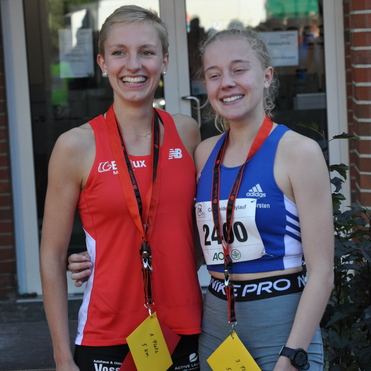 Zwei junge Sportlerinnen posieren lächelnd nach einem Wettkampf, jeweils mit Medaillen und Platzierungs-Schildern.