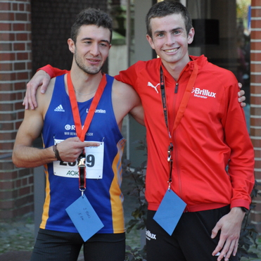 Zwei Athleten posieren l&auml;chelnd nach einem Wettkampf, beide tragen Medaillen und Wettkampfnummern.