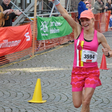 L&auml;uferin in pinkem Outfit jubelt mit zwei Fingern, w&auml;hrend sie die Ziellinie &uuml;berquert. Zuschauer applaudieren im Hintergrund.