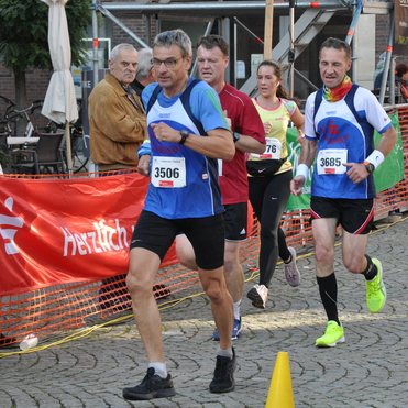 Vier L&auml;ufer in Sportkleidung w&auml;hrend eines Wettbewerbs, umgeben von Zuschauer- und Veranstaltungs&uuml;berst&auml;nden.