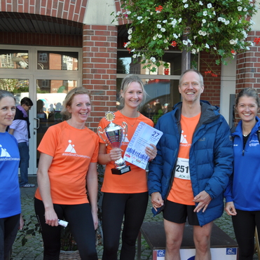 Gruppe von f&uuml;nf Personen in Laufbekleidung, eine Person h&auml;lt einen Pokal und eine Urkunde vor einer Backsteinmauer.