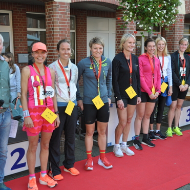 Mehrere L&auml;uferinnen stehen auf einem Podium mit Medaillen und Urkunden, w&auml;hrend ein Moderator mit Mikrofon spricht.