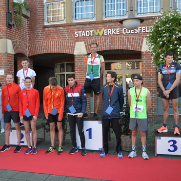 Siegerehrung mit f&uuml;nf L&auml;ufern auf Podesten, einem Moderator und Zuschauern vor einer Ziegelsteinfassade.