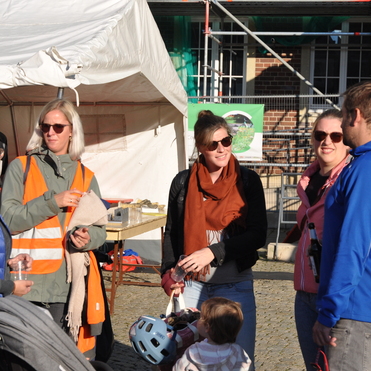 Sechs Personen stehen bei einem Fest, im Hintergrund ein Zelt und eine Baustelle, eine Frau h&auml;lt ein Kind auf dem Arm.