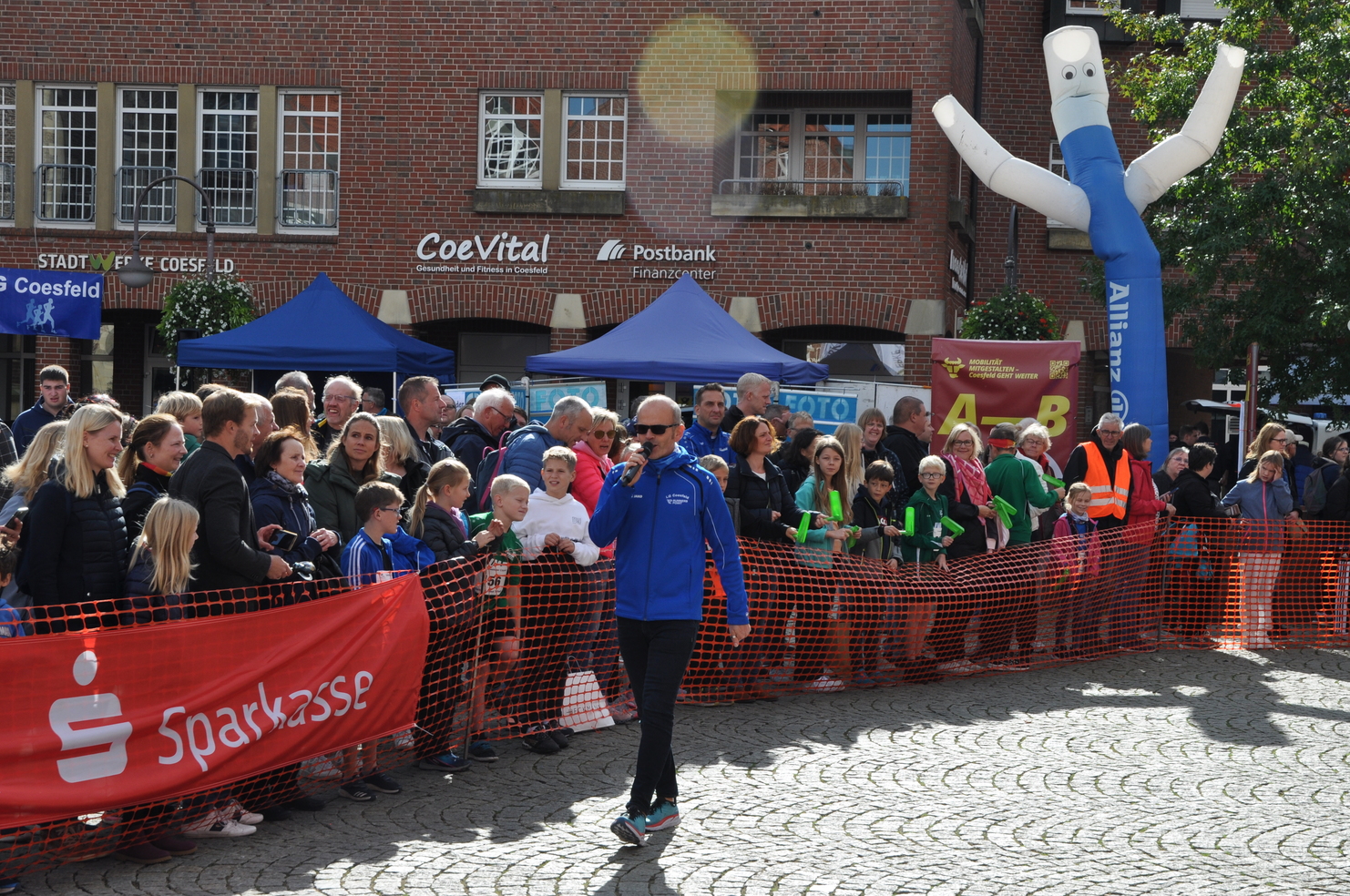 Ein Moderator in blauer Jacke spricht mit Publikum, umgeben von Zuschauern und bunten Ständen auf einem Fest.