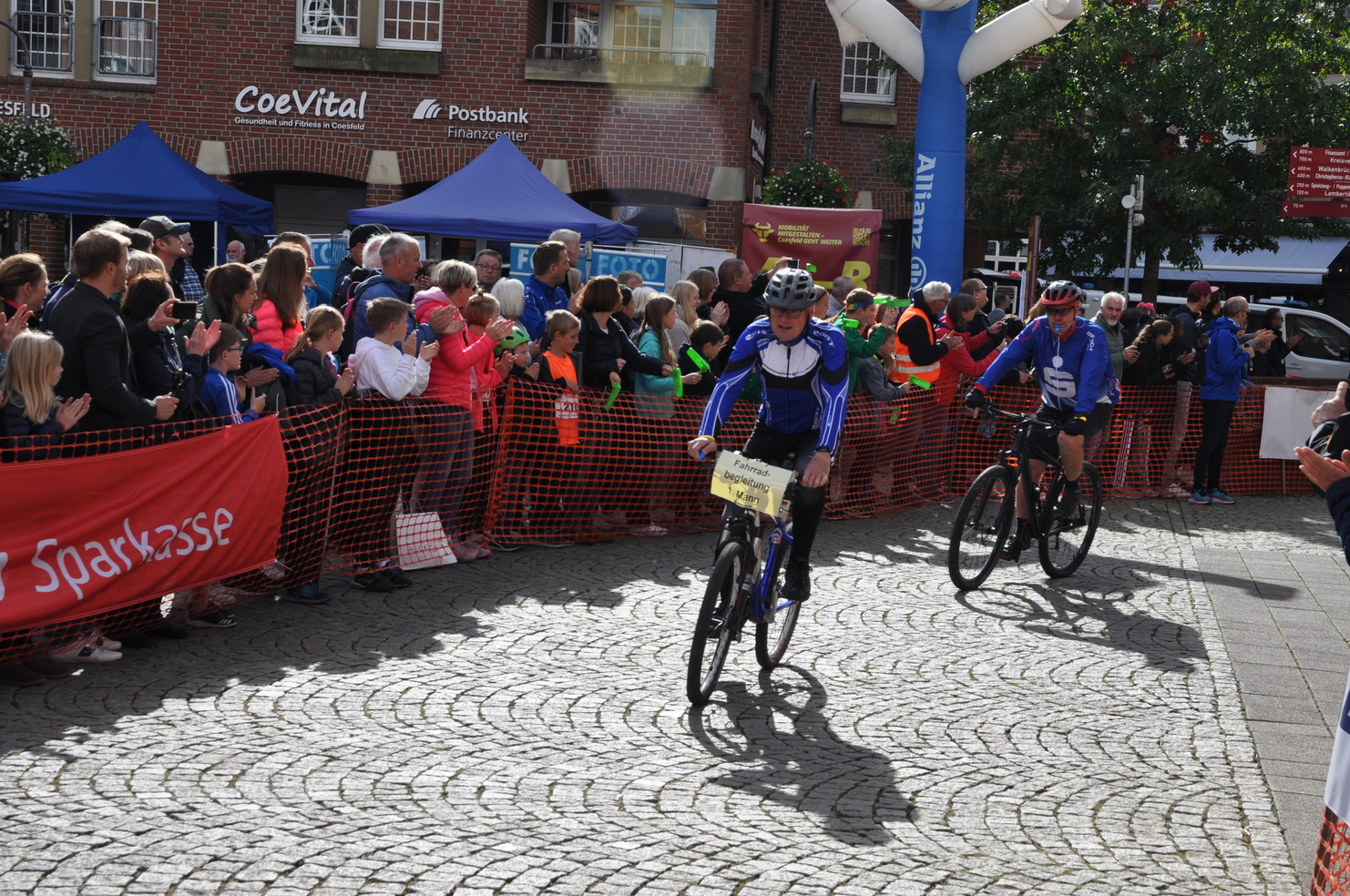 Zwei Radfahrer auf einer gepflasterten Straße fahren einem jubelnden Publikum entgegen, während sie ein Rennen beenden.