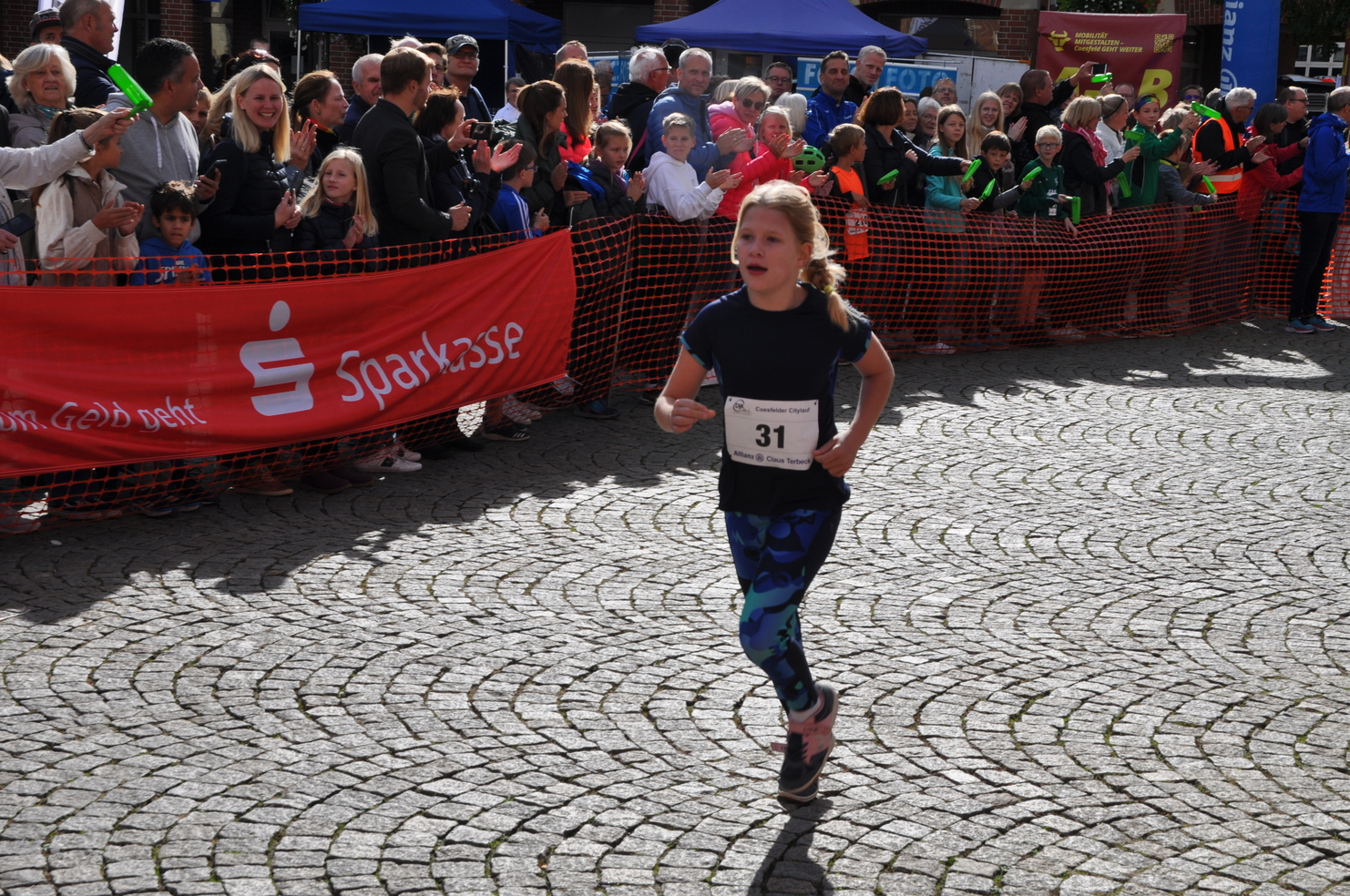 Mädchen mit Startnummer 31 läuft auf einem Wettkampf, umgeben von jubelnden Zuschauern und einer roten Werbebande.