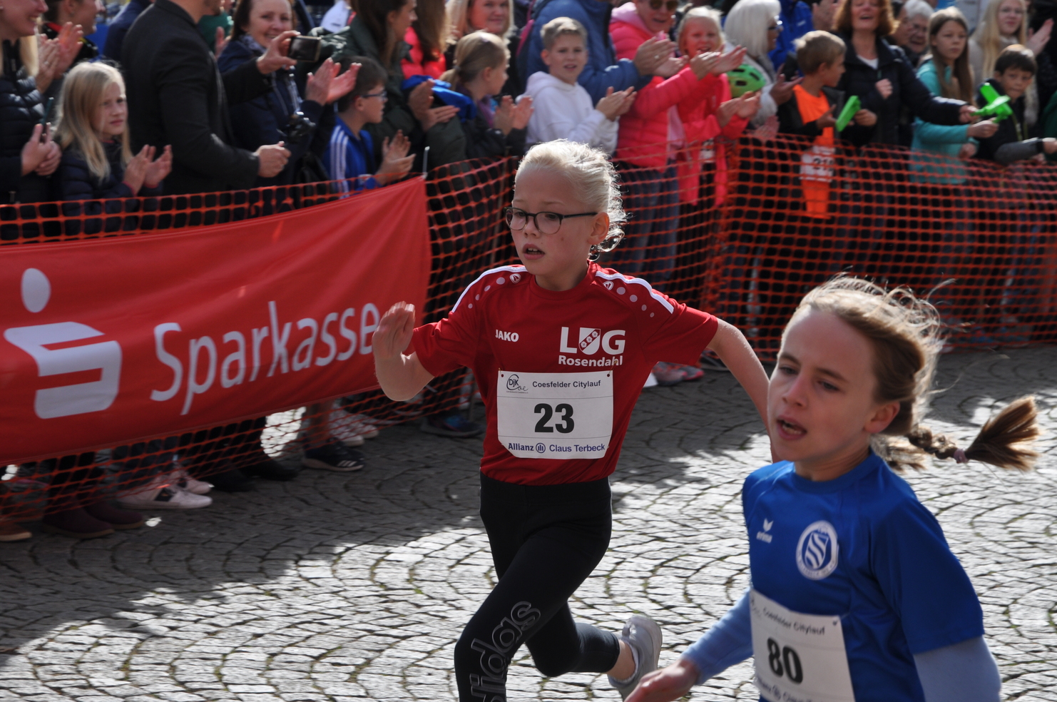 Zwei Kinder in Sportkleidung laufen beim Coesfelder Citylauf, viele Zuschauer stehen applaudierend im Hintergrund.