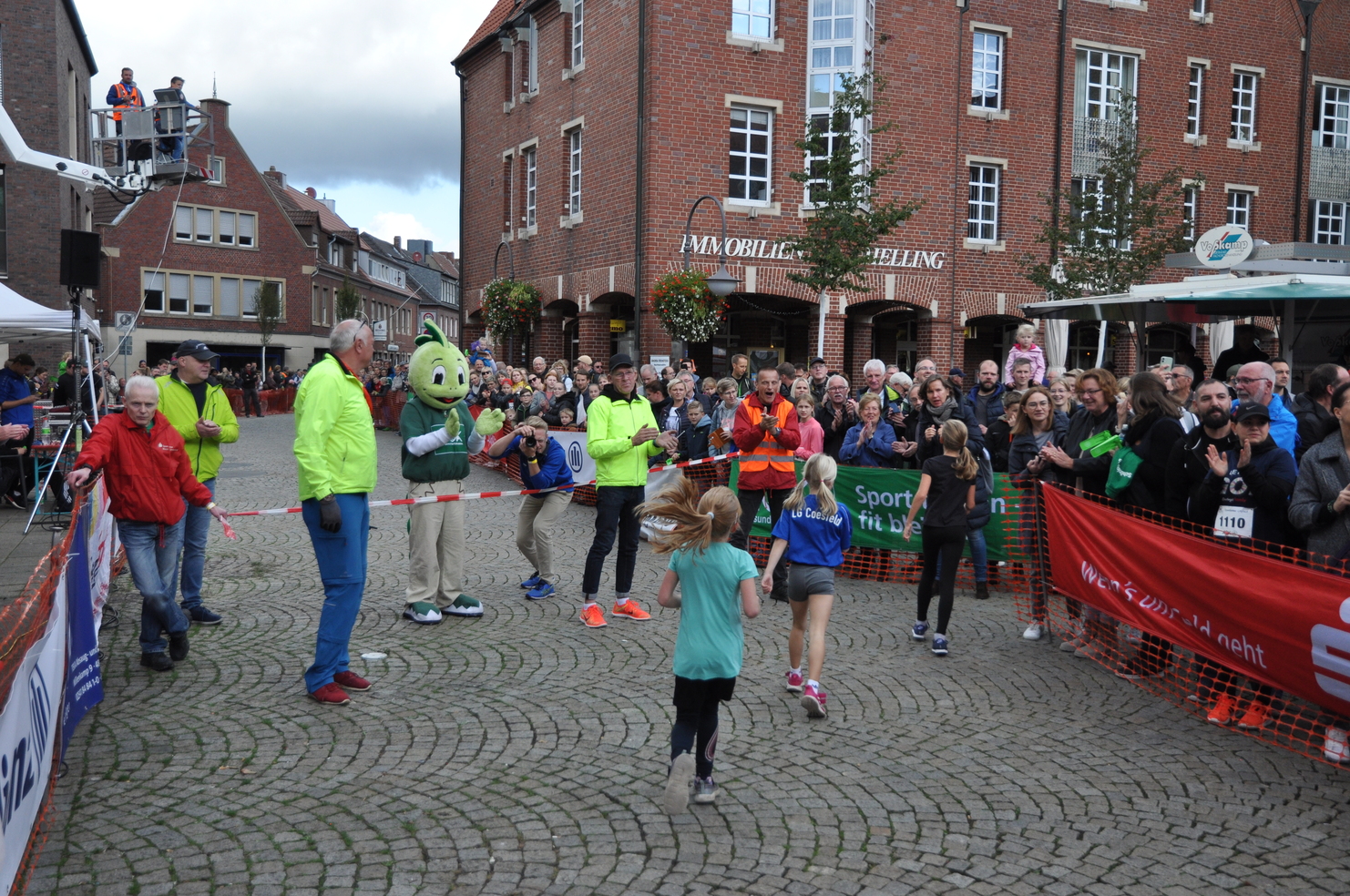 Zwei Kinder laufen im Zielbereich eines Wettlaufs, umgeben von jubelnden Zuschauern und Helfern in Neonkleidung.