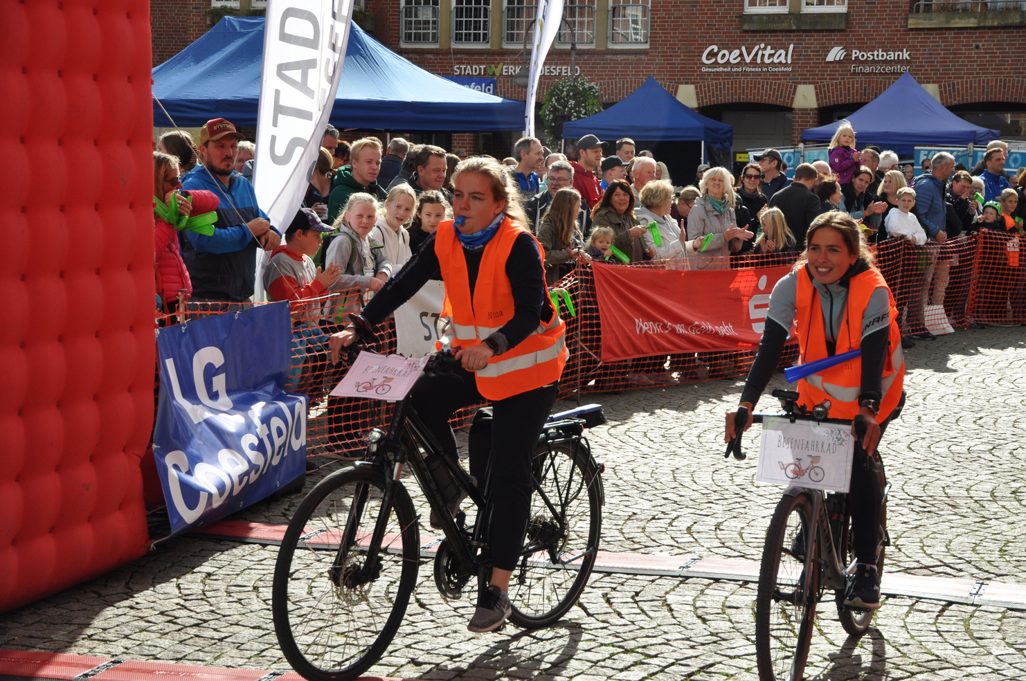 Zwei Radfahrerinnen in auffälligen Westen fahren durch das Ziel eines Radrennens, umgeben von jubelndem Publikum.