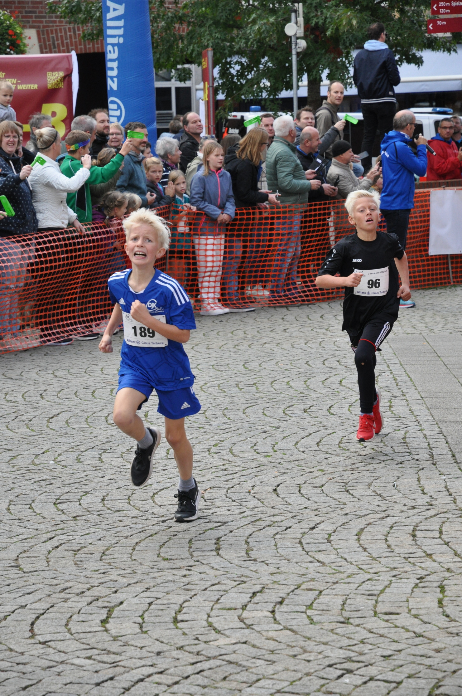 Zwei Kinder im Wettlauf, eines in blauem und das andere in schwarzem Trikot, laufen auf einem gepflasterten Platz.
