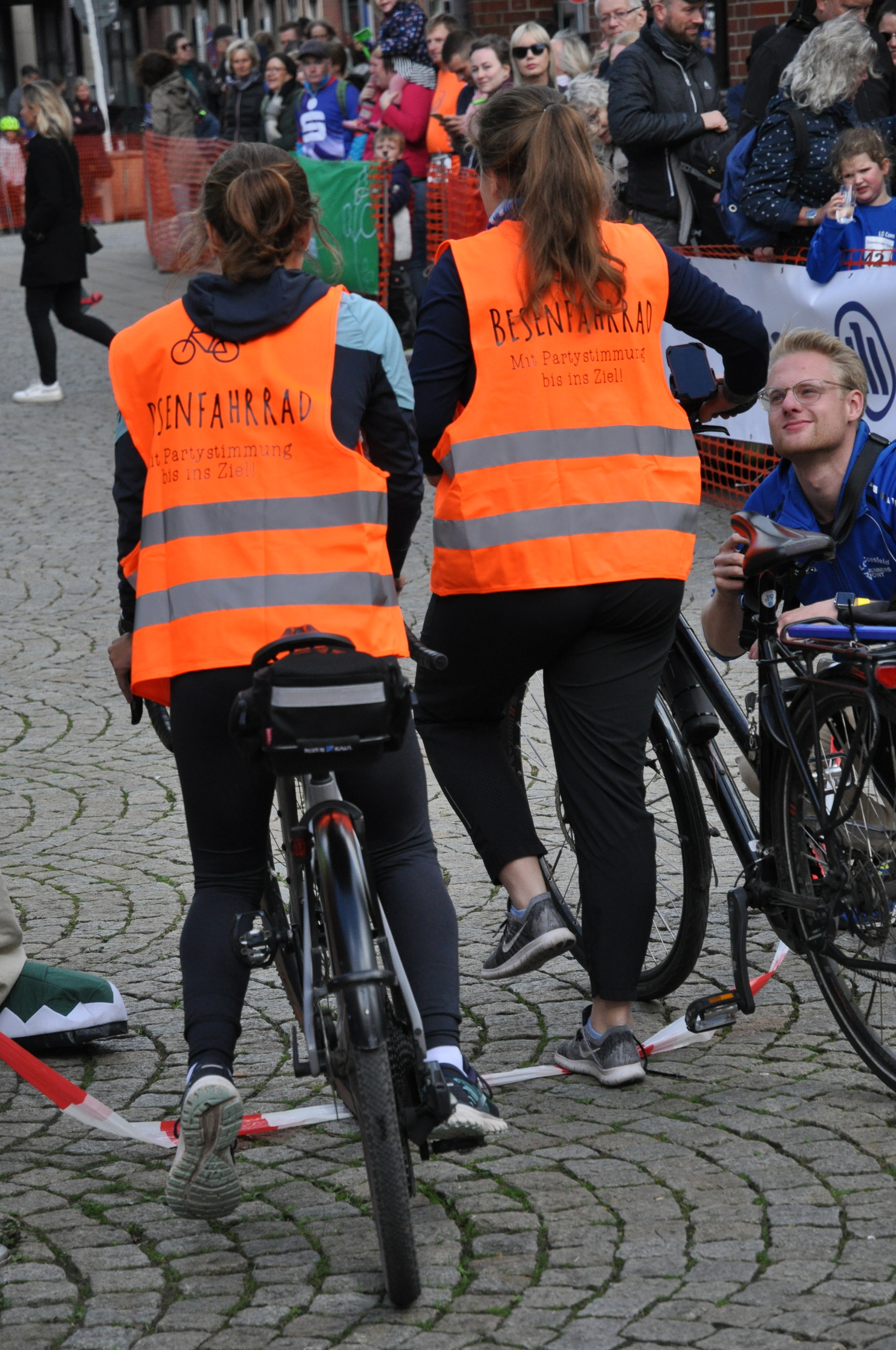 Zwei Personen in leuchtend orangefarbenen Westen stehen mit Fahrrädern vor einer Menschenmenge auf einem Platz.