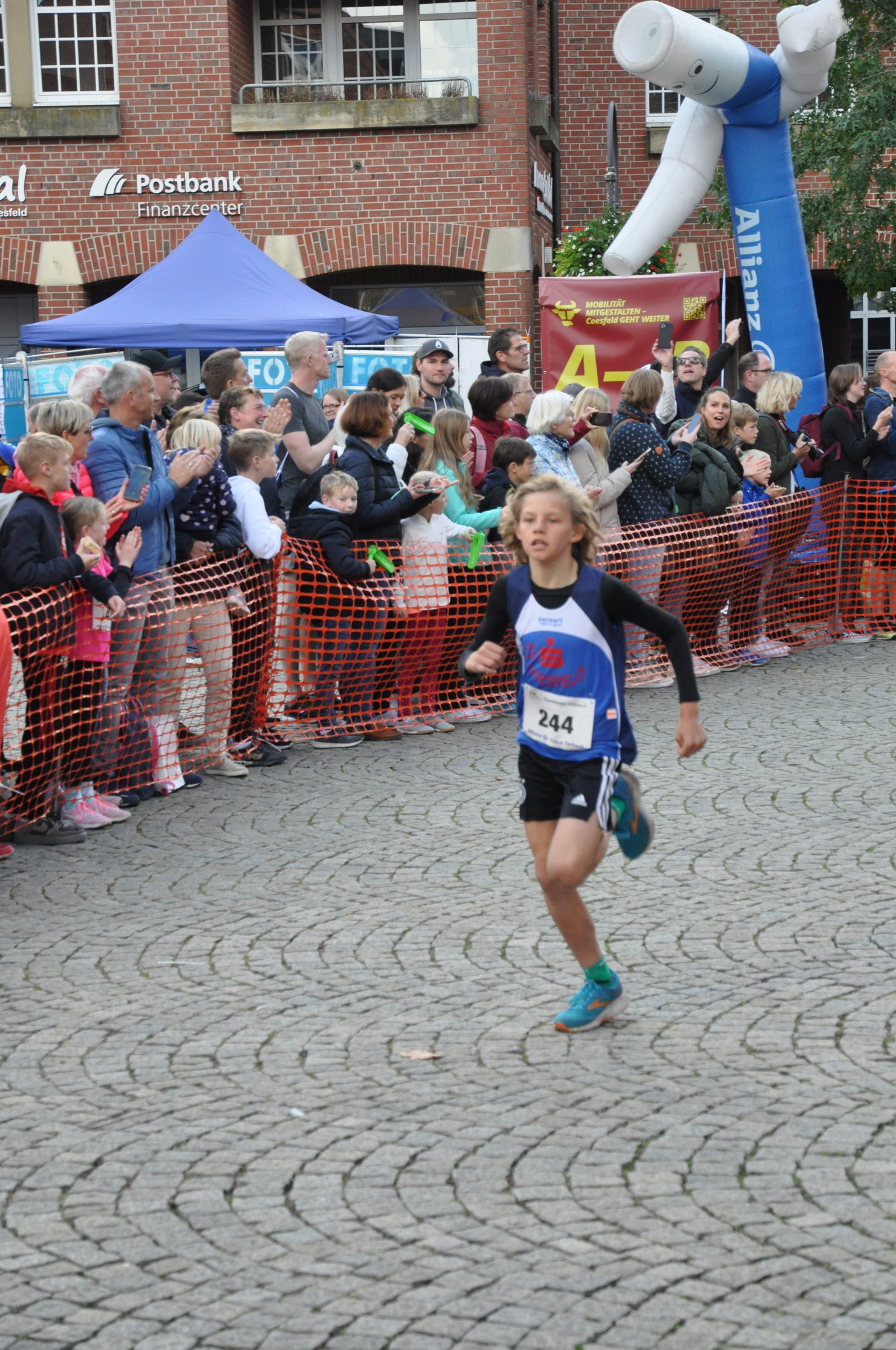 Junger Läufer in Sportbekleidung rennt auf einer gepflasterten Straße, Zuschauer feuern ihn an.