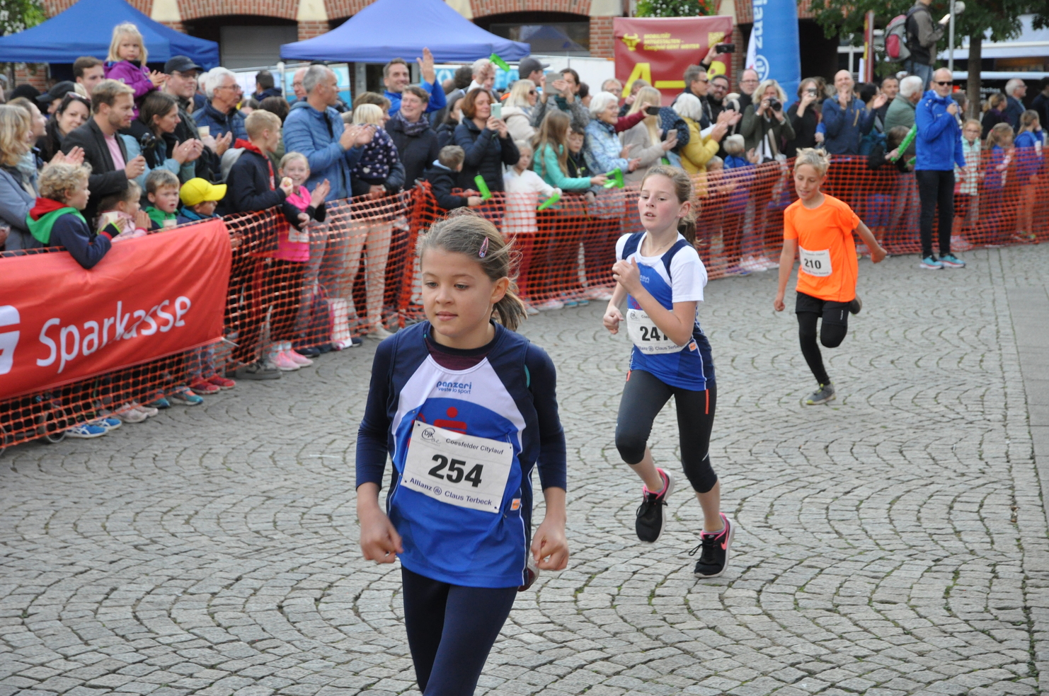 Drei Kinder laufen beim Wettlauf, umgeben von jubelnden Zuschauern und einem roten Sparkassen-Banner.