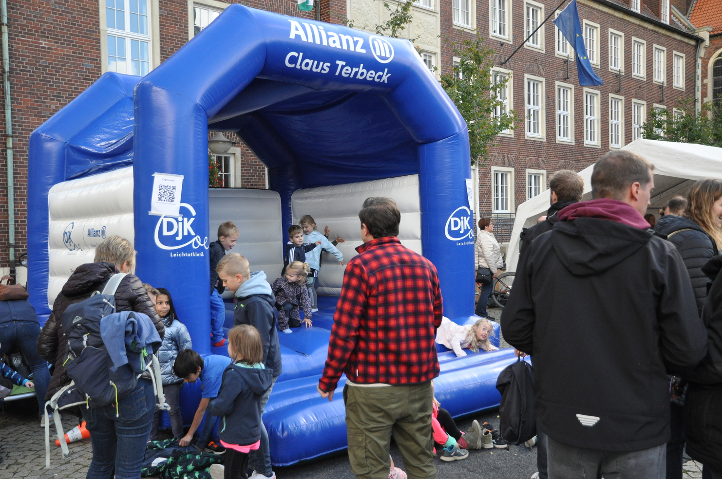 Kinder spielen auf einer blauen Hüpfburg, umgeben von Erwachsenen, in einem belebten Stadtbereich.