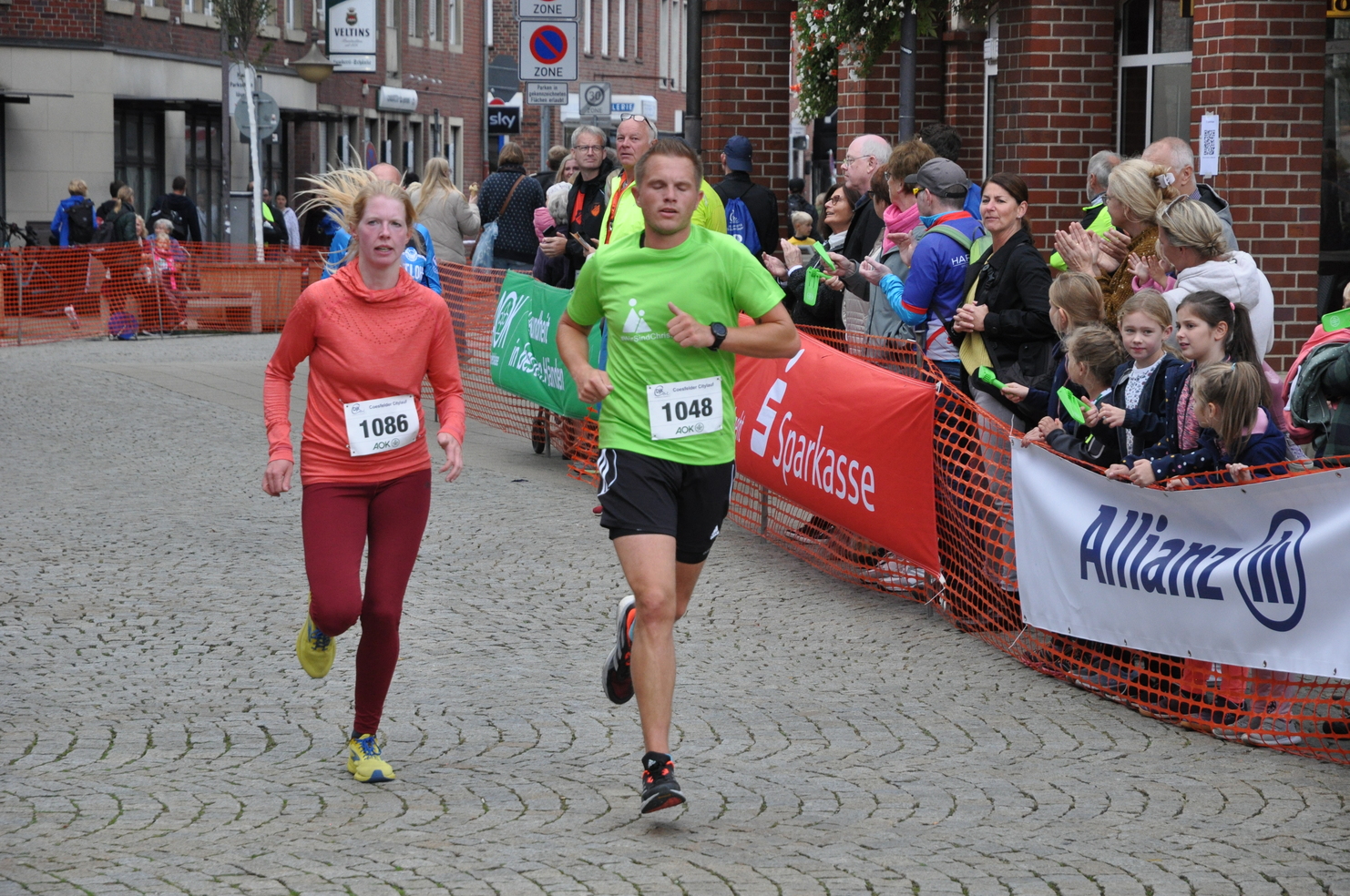 Zwei Läufer, einer in Grün und einer in Rot, laufen auf einer Straße, umgeben von jubelnden Zuschauern.
