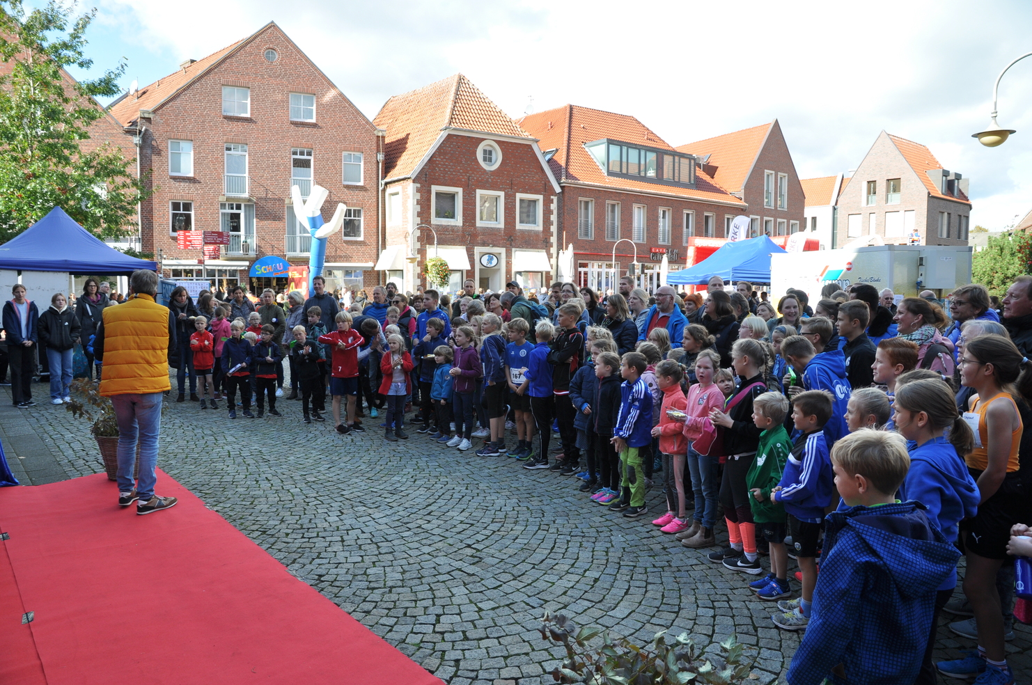 Gruppierung von Kindern und Erwachsenen bei einer Veranstaltung auf einem Platz, Blick auf eine Bühne im Vordergrund.